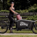 Een persoon met sportkleding en een pet zit glimlachend op een zwarte bakfiets met het opschrift "De Gym Koerier". De fiets heeft een grote voorbak met daarop een yogamat, met groen en houten traptreden op de achtergrond.