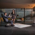 Een man in pak zit op een bureaustoel van autobanden en houdt een mok vast, terwijl een vrouw in een badjas koffie voor hem inschenkt in een moderne woonkamer met grote ramen. Buiten is een auto zichtbaar door het glas.