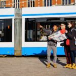 Drie mensen staan voor een blauwwitte tram en houden een kaart vast. Eén persoon wijst vooruit terwijl de anderen in dezelfde richting kijken, alsof ze navigeren of bezienswaardigheden bekijken in een stedelijke omgeving.