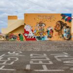 Een geel gebouw heeft kleurrijke muurschilderingen met abstracte gezichten, ogen en dieren. Op de stoep ervoor staan witgeschilderde letters met vakantiegroeten in het Portugees. De lucht is gedeeltelijk bewolkt.