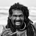 Een man met lange dreadlocks lacht breed, laat zijn tanden zien en maakt een "shaka"-handgebaar. Hij lijkt buiten te zijn, mogelijk op een strand, met golven zichtbaar in de wazige achtergrond. De foto is in zwart-wit.