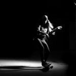 Een persoon met een pet op en casual kleding skateboardt onder een dramatische spot, omgeven door duisternis, die een lange schaduw op de vloer werpt.