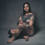 Een vrouw met lang donker haar zit op de grond tegen een grijze achtergrond, naakt, met armen en benen gekruist. Haar lichaam is bedekt met kleurrijke tatoeages, waaronder grote vleugels op haar borst en verschillende ontwerpen op haar armen en benen.