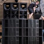 Twee mensen zetten buiten een grote stapel zwarte luidsprekers en subwoofers op, waarbij één persoon op de luidsprekers staat en de ander de apparatuur aanreikt. Op de achtergrond zijn bomen en tuigage zichtbaar.