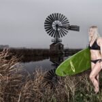 Een vrouw in een zwarte bikini houdt een groene surfplank vast terwijl ze bij een met gras begroeide rivieroever staat, met een grote windmolen en een bewolkte lucht op de achtergrond.
