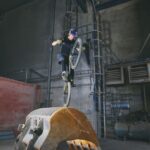 Een fietser met veiligheidsuitrusting maakt een wheelie bovenop een grote, roestige graafbak in een industrieel magazijn met betonnen muren en industriële apparatuur.