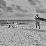 Zwart-witfoto van een strandtafereel met verschillende mensen; één persoon staat een handdoek uit te schudden, anderen zitten of lopen langs de kust, met zachte golven en wolken in de lucht.