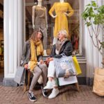 Twee vrouwen zitten op een bankje voor een kledingwinkel, lachen en houden boodschappentassen vast. Achter hen in de etalage staan etalagepoppen in stijlvolle gele outfits. Naast het bankje staat een potplant.