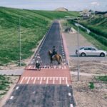 Een fietser en een ruiter kruisen elkaar op een verharde weg met groene grasbermen aan weerszijden. Een geparkeerde auto en een kanaal zijn zichtbaar aan de rechterkant, met huizen op de achtergrond onder een gedeeltelijk bewolkte hemel.