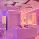 Een moderne, betegelde badkamer wordt verlicht door roze en paarse LED-lampen, met een centrale eilandwastafel, spiegels, toiletartikelen, een metalen kar vol toiletpapier en een minimalistisch, strak design.