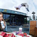 Een jonge vrouw drinkt frisdrank en eet frietjes aan een McDonald's tafel buiten, terwijl op de achtergrond iemand een backflip maakt in de lucht bij de ingang van het restaurant.