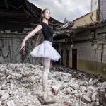 Een jonge ballerina in een zwarte maillot en witte tutu poseert en pointe tussen het puin van een verwoest gebouw, waarbij elegantie en kwetsbaarheid in contrast staan met de verwoesting om haar heen.