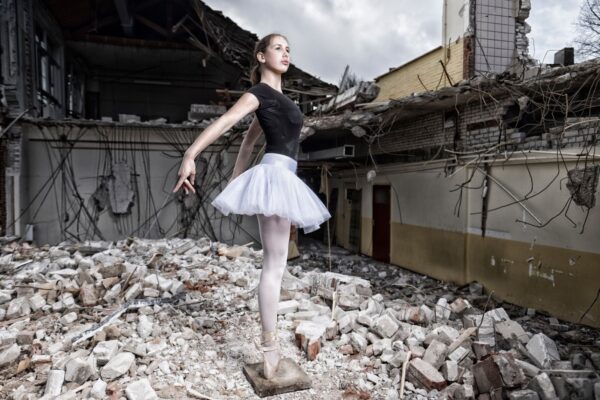 Een jonge ballerina in een zwarte maillot en witte tutu poseert en pointe tussen het puin van een verwoest gebouw, waarbij elegantie en kwetsbaarheid in contrast staan met de verwoesting om haar heen.