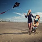 Een vrouw in een wetsuit staat op blote voeten op een zandstrand, houdt een kiteboard vast en vliegt op een zwarte vlieger met een schedel en gekruiste botten ontwerp. De lucht is gedeeltelijk bewolkt en op de achtergrond is de oceaan zichtbaar.