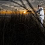 Een honkbalspeler in een wit uniform en pet staat in het hoge helmgras, houdt een knuppel vast alsof hij klaar is om te slaan, met achter hem de zon die ondergaat aan de horizon en dramatische wolken in de lucht.
