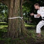Een persoon in een wit judo-uniform kijkt naar een grote boom met een witte riem om de stam in een park. Hij lijkt te oefenen of zich voor te bereiden op een vechtsport. Zonlicht valt door de omringende bomen.