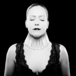 Een vrouw met gesloten ogen en donkere lippenstift draagt een decoratief, verticaal halsstuk en een met kant afgezet zwart topje en poseert tegen een effen zwarte achtergrond op een contrastrijke zwart-witfoto.