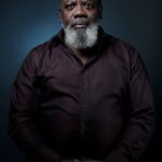 Een man van middelbare leeftijd met een grijze baard en kort haar zit tegen een donkere achtergrond, draagt een donker kastanjebruin overhemd en een lichtgekleurde broek en kijkt kalm naar de camera met zijn handen rustend op zijn schoot.