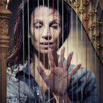 Een vrouw in een spijkerblouse staat achter een harp, met opgeheven hand en gespreide vingers. De snaren van de harp zijn zichtbaar op de voorgrond, waardoor haar gezicht gedeeltelijk aan het oog onttrokken wordt.