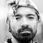 Een zwart-wit close-up portret van een man met een baard, die recht in de camera kijkt. Hij draagt een hoofddoek met patroon rond zijn hoofd. De achtergrond is onscherp.
