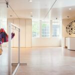 Een etalagepop met een zwarte hoed, jas en rok met patroon staat naast kleurrijke sjaals in een lichte, moderne winkel met grote ramen en een muurschildering van een boom bij de kassa.