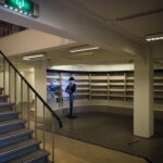 Een grotendeels lege, moderne boekwinkel of bibliotheek met kale planken. Aan de linkerkant is een trap en een etalagepop met hoed en jas staat bij een kleine boekenkast in de hoek. Bovenlichten verlichten de ruimte.