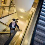 Een persoon met een hoed en sjaal loopt op een roltrap naar boven in een modern gebouw, met een lege roltrap naar beneden naast hem en een bordje "Welkom" zichtbaar op de muur voor hem.