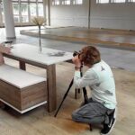 Een persoon knielt terwijl hij een etalagepop op kinderformaat fotografeert, gekleed in een hoed en jas, zittend aan een moderne tafel in een ruime, lichte kamer met grote ramen en houten vloeren.