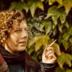 Een persoon met krullend haar en een sjaal met patroon houdt een brandende sigaret vast en ademt rook uit. Hij staat voor een muur bedekt met groene bladeren.