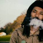 Een persoon met lange dreadlocks en een muts ademt buiten een rookwolk uit, met bomen en groen wazig op de achtergrond.