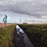 Een vrouw in een turquoise shirt draagt een surfplank langs een landelijk pad naast een smal waterkanaal, met windturbines op de achtergrond onder een bewolkte hemel.