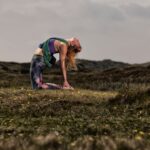 Een vrouw beoefent yoga in de buitenlucht op een grasveld, waarbij ze een kamelenhouding aanneemt met haar hoofd naar achteren gekanteld en haar handen op haar hielen, omringd door heuvels onder een bewolkte hemel.