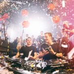Twee DJ's treden op in een levendige club, omringd door confetti en ballonnen, terwijl een menigte geniet van de muziek en fotografen de energieke sfeer vastleggen.