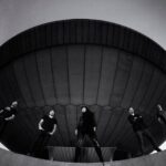 Een zwart-witfoto van vijf mensen die in een brede halve cirkel onder een groot, koepelvormig bouwwerk staan, gezien vanuit een lage hoek tegen de lucht.