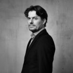 Een man met golvend donker haar en lichte stoppels, draagt een donker colbert en licht overhemd, staat in profiel tegen een effen grijze achtergrond met textuur en kijkt serieus naar de camera. De afbeelding is in zwart-wit.