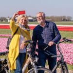 Een ouder echtpaar met fietsen neemt samen een selfie voor levendige rijen bloeiende tulpenvelden op een zonnige dag. De vrouw draagt een gele jas en de man een marineblauwe jas. Beiden glimlachen en genieten van het moment.