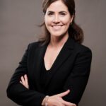 Een vrouw met donker haar en een zwarte blazer staat tegen een effen grijze achtergrond en glimlacht zelfverzekerd met haar armen over elkaar.