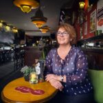 Een glimlachende vrouw met bril zit aan een ronde tafel in een gezellige pub, versierd met posters, planten en warme verlichting. De kroeg lijkt leeg behalve zij, wat een uitnodigende sfeer creëert.