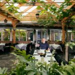 Een man zit alleen aan een tafel in een zonovergoten restaurant met een houten pergola, bladerrijke planten en geruite tafelkleden, en heft het glas in een ruime, groene eethoek.