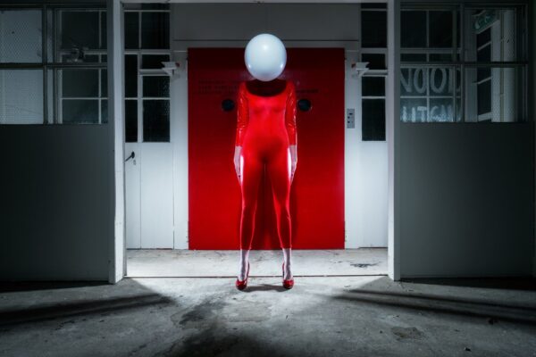 Een persoon in een glimmend rood lijfje en hoge hakken staat naar voren te kijken. Hun hoofd is bedekt met een grote witte bol. Ze staan in een zwak verlichte industriële ruimte met witte muren en een felrode deur achter hen.