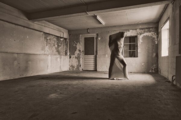 Een persoon staat in een lege, vervallen kamer, strak ingepakt in een laken dat zijn gelaatstrekken verhult. Licht van een raam werpt schaduwen op de versleten vloer en de afbladderende muren, wat een dramatische en griezelige sfeer creëert.