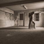 Een persoon staat in het midden van een lege, versleten kamer, strak gewikkeld in een dunne, doorschijnende doek die zijn gelaatstrekken verhult. Zonlicht stroomt door een raam en werpt schaduwen op de gestructureerde muren en vloer.