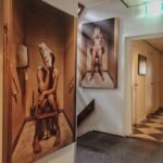 Grote muurschilderingen in een gang tonen naaktfiguren die op een toilet zitten, de een met een krant over zijn gezicht en de ander met een voorwerp dat zijn hoofd bedekt. De gang heeft tapijt en tegelvloeren, witte muren en verschillende deuren.