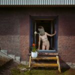 Een naakt persoon staat voor een raam en houdt een gieter boven zijn hoofd terwijl het water naar buiten stroomt. Een potplant staat op een klein houten platform onder het raam, bevestigd aan een bakstenen gebouw.