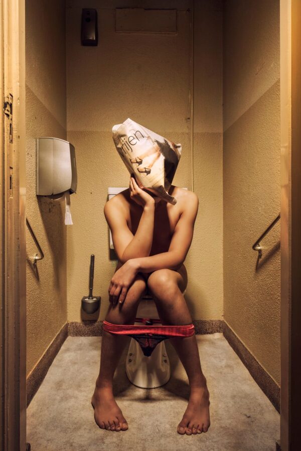 Een persoon zit op een toilet in een schemerig toilet, met alleen ondergoed aan. Ze houden een papieren zak met een gezicht erop boven hun hoofd.