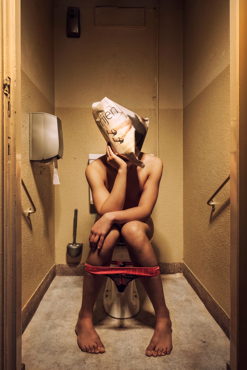 Een persoon zit op een toilet in een schemerig toilet, met alleen ondergoed aan. Ze houden een papieren zak met een gezicht erop boven hun hoofd.