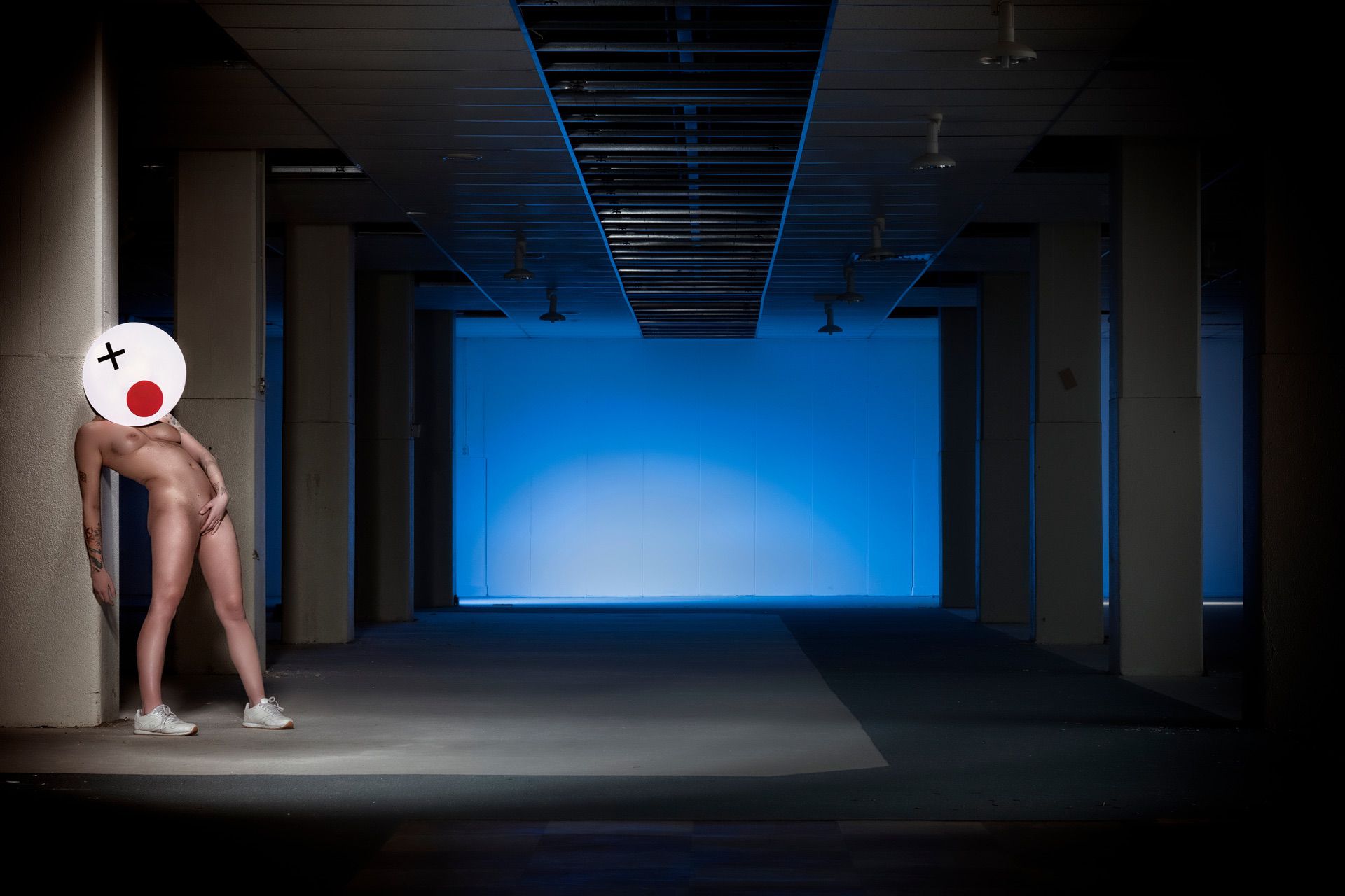 Een naakt persoon met witte gympen en een masker met een cartoongezicht leunt tegen een zuil in een schemerige, lege kamer met blauwe verlichting op de achtergrond.