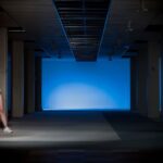 Een naakt persoon met witte gympen staat tegen een pilaar in een schemerige, lege kamer met zuilen en een blauw verlichte achtergrond. Hun gezicht is bedekt met een cartoonesk, rond masker met een verbaasde uitdrukking.