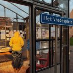 Een vrouw met lang blond haar staat in een glazen constructie en bekijkt displayborden. Een blauw bord met "Het Vredesplein" is zichtbaar op de deur en het interieur heeft een tapijt en meubels.