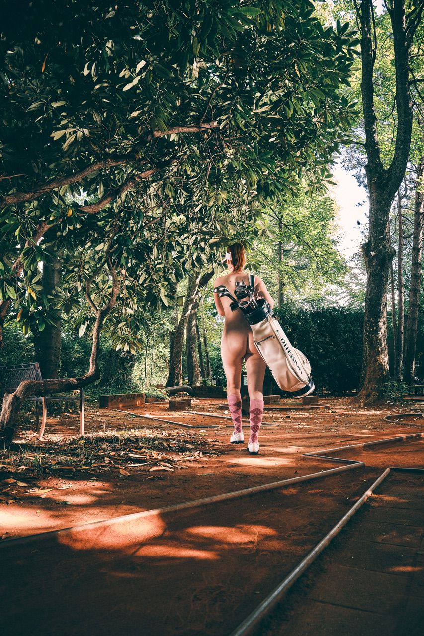 Een persoon, van achteren gezien, loopt over een schaduwrijk buitenpad met een grote golftas. Ze dragen alleen sokken en schoenen, omringd door bomen en groen.